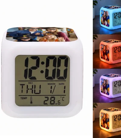 A studio product shot of the Upahar18 personalized photo printed led alarm clock in a square cube shape. The front-facing digital screen clearly displays a black numeric font showing the time as 12:00, day as THU, month and date as 1/1, and temperature as 28.6°C. The top surface features a colorful family photo printed graphic of parents and children in Halloween costumes. On the right side, a vertical stack of four smaller images displays the clock glowing on a wooden table in blue, red, purple, and yellow ambient nightlight modes, illuminating a custom photo printed panel of a family on its side.